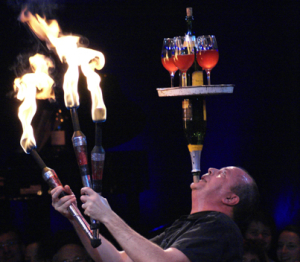 Steve juggling fire clubs, while balancing bottles and glasses on his chin.